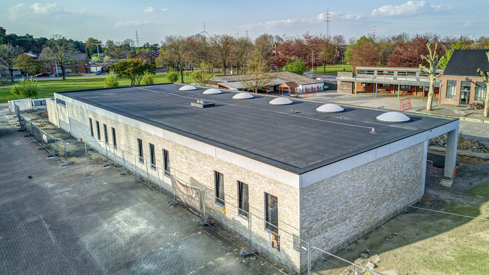 Aerial view of a brick building with a flat roof and multiple rounded skylights, surrounded by trees and a playground area.