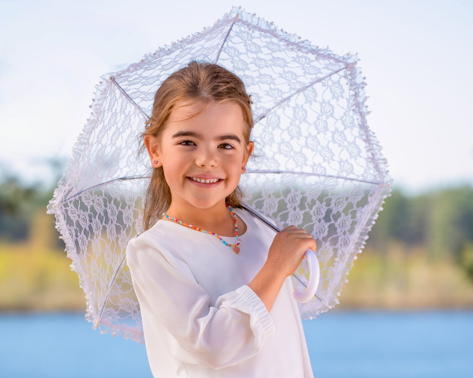Smiling girl in a white blouse and coral skirt holding a lace parasol at the beach by the water.