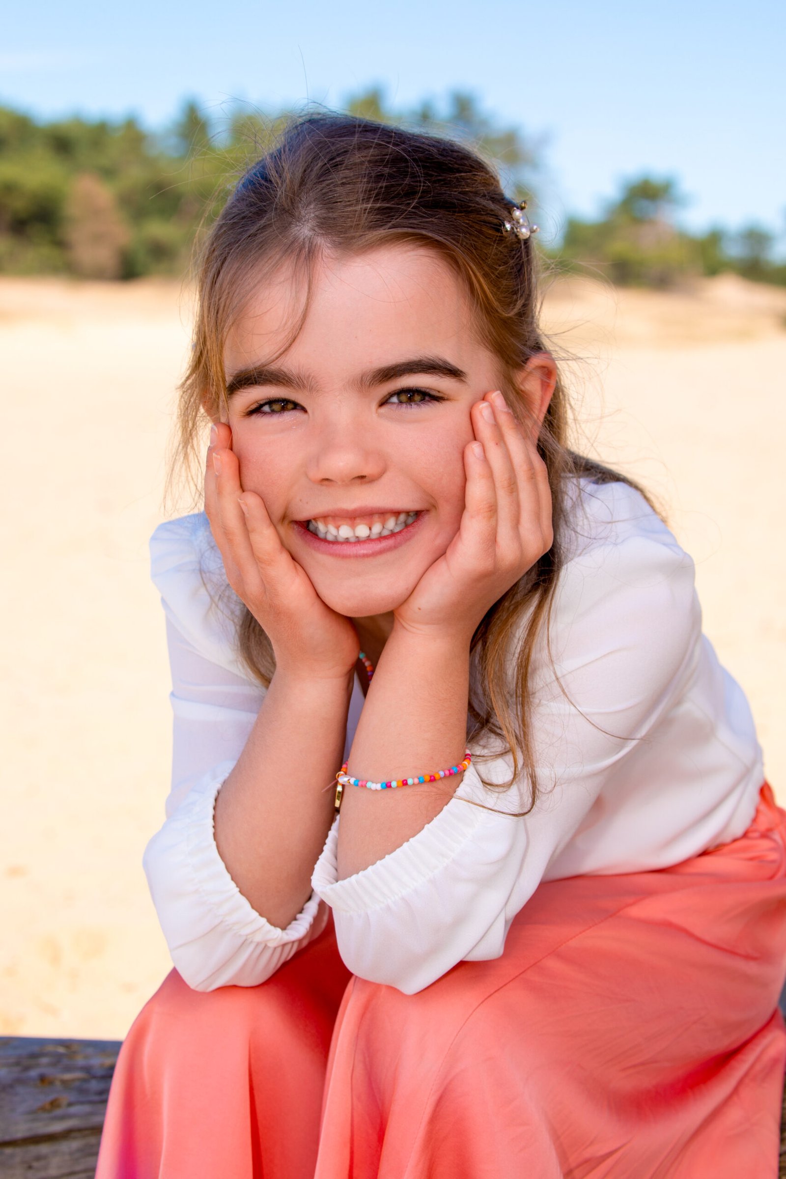 Girl lying on a pink blanket outdoors, smiling with hands pressed to her cheeks, wearing a white blouse and pink skirt.