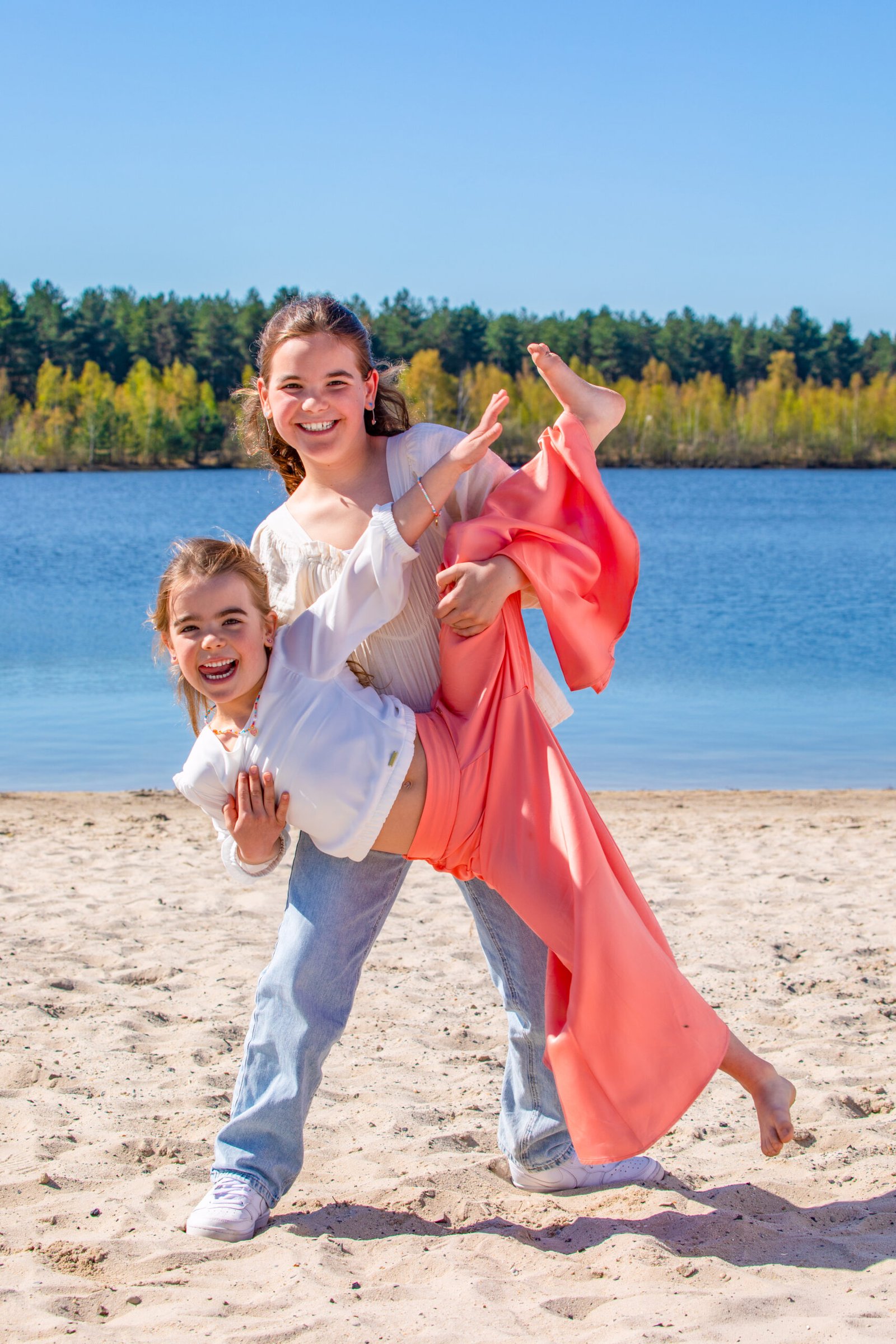 Two smiling girls play on a sandy beach by a calm blue lake with a forest in the background censored by trees.