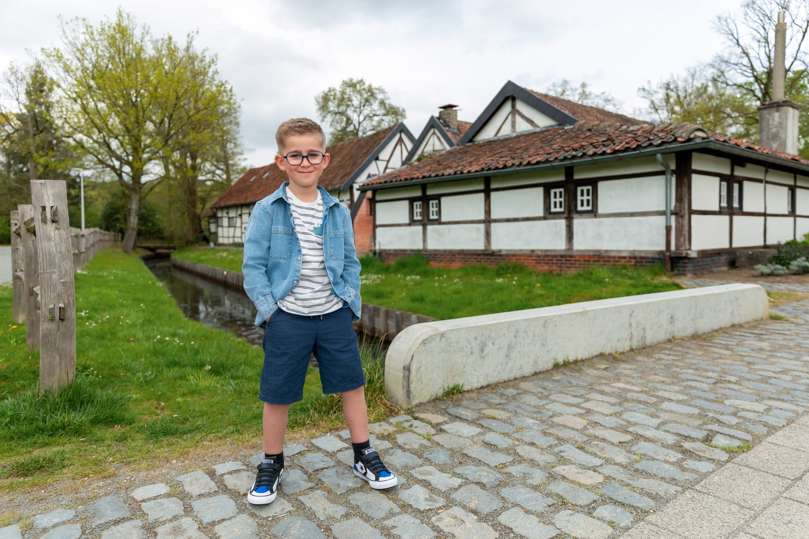 Boy in a denim jacket and striped shirt poses on a cobblestone path beside a canal with white timber-framed houses in the background.