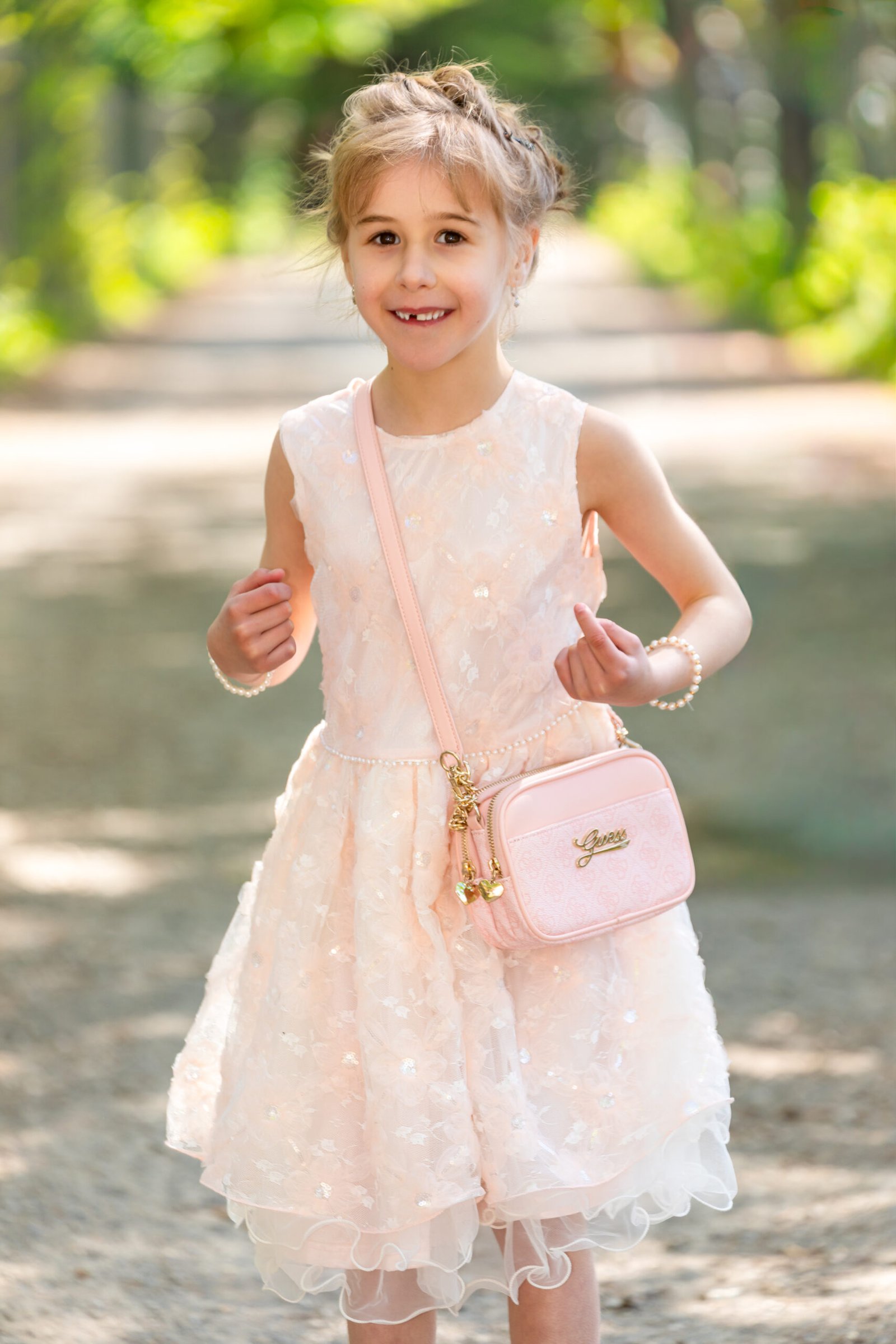 Young girl in a pale pink lace dress with a matching pink crossbody bag, smiling on a sunlit park path.