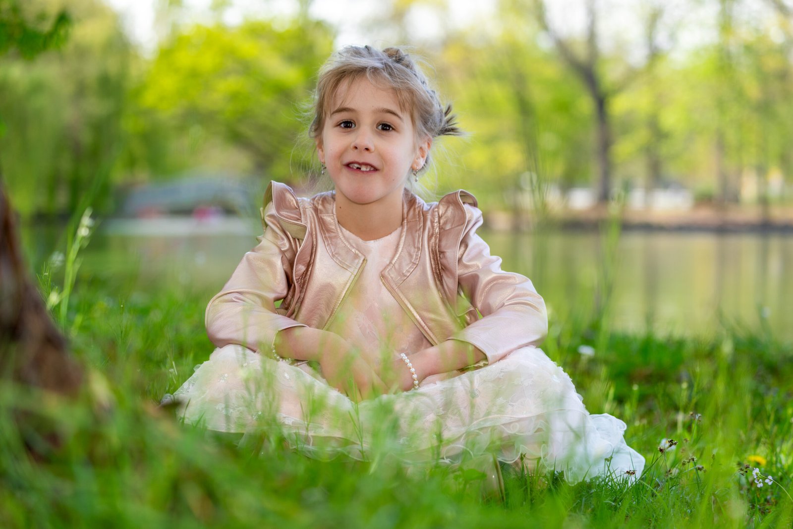 Smiling young girl in a pink dress sits on grass in a sunny park with blurred trees in the background.",