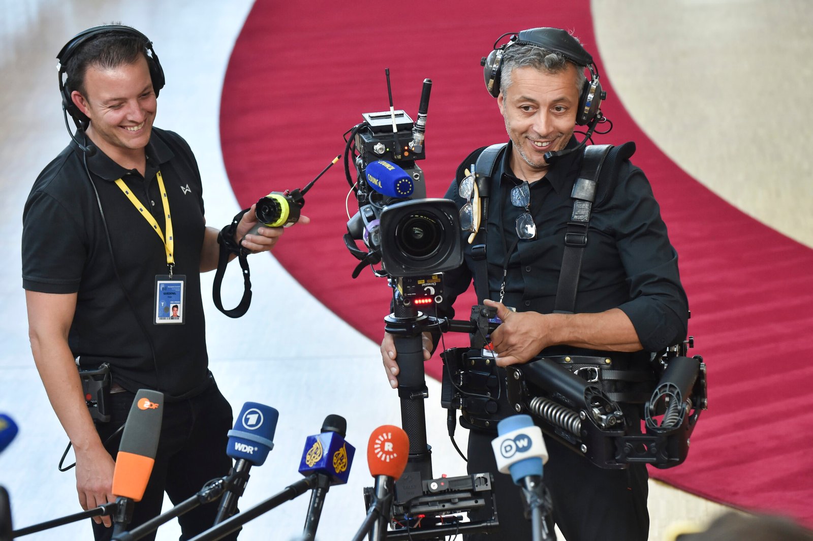 Two videographers in black shirts at a press conference, smiling, with a large camera rig and multiple microphones in front of them.