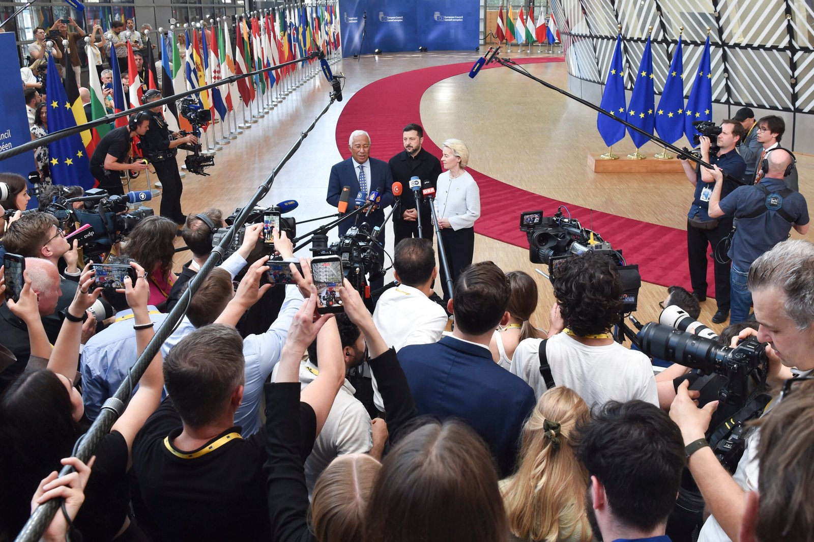 Three speakers addressing a press conference, surrounded by cameras and reporters in a flag-filled hall.