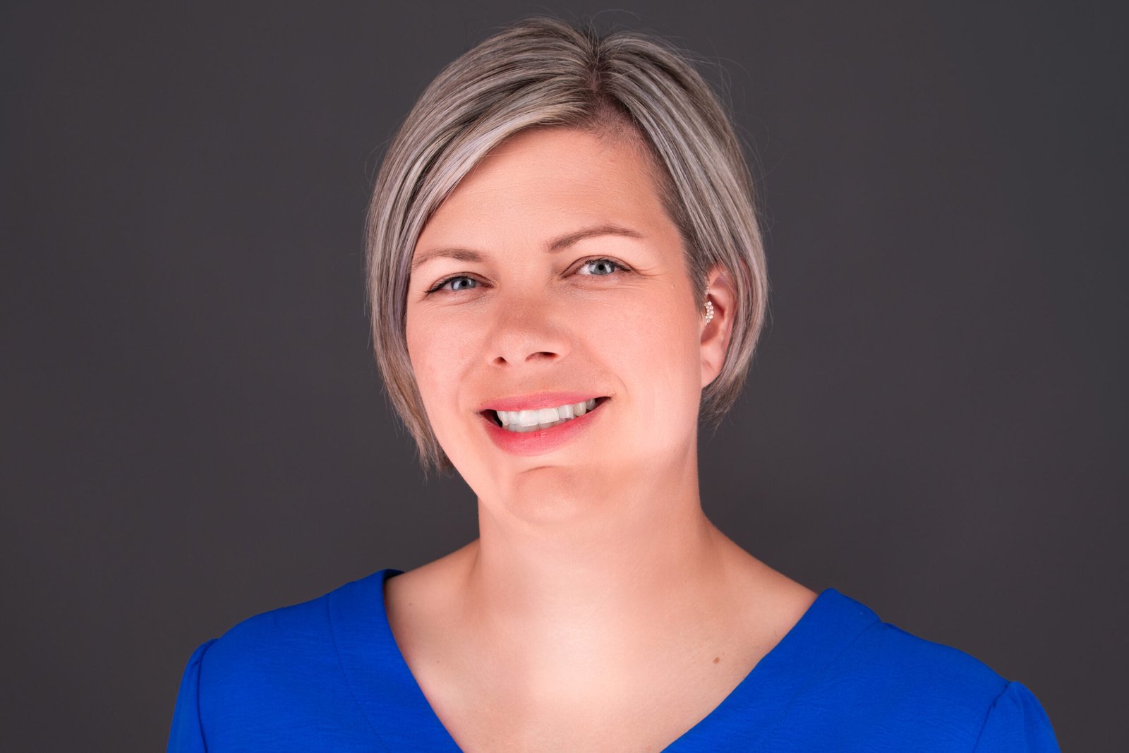 Smiling woman with short gray-blonde hair wearing a blue top against a dark background.