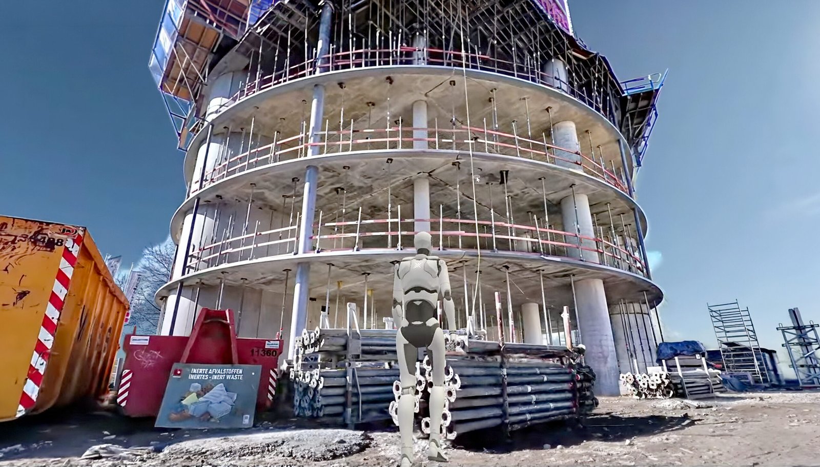 Under-construction circular concrete tower with metal scaffolding and safety barriers; a life-size humanoid robot statue stands in the foreground.